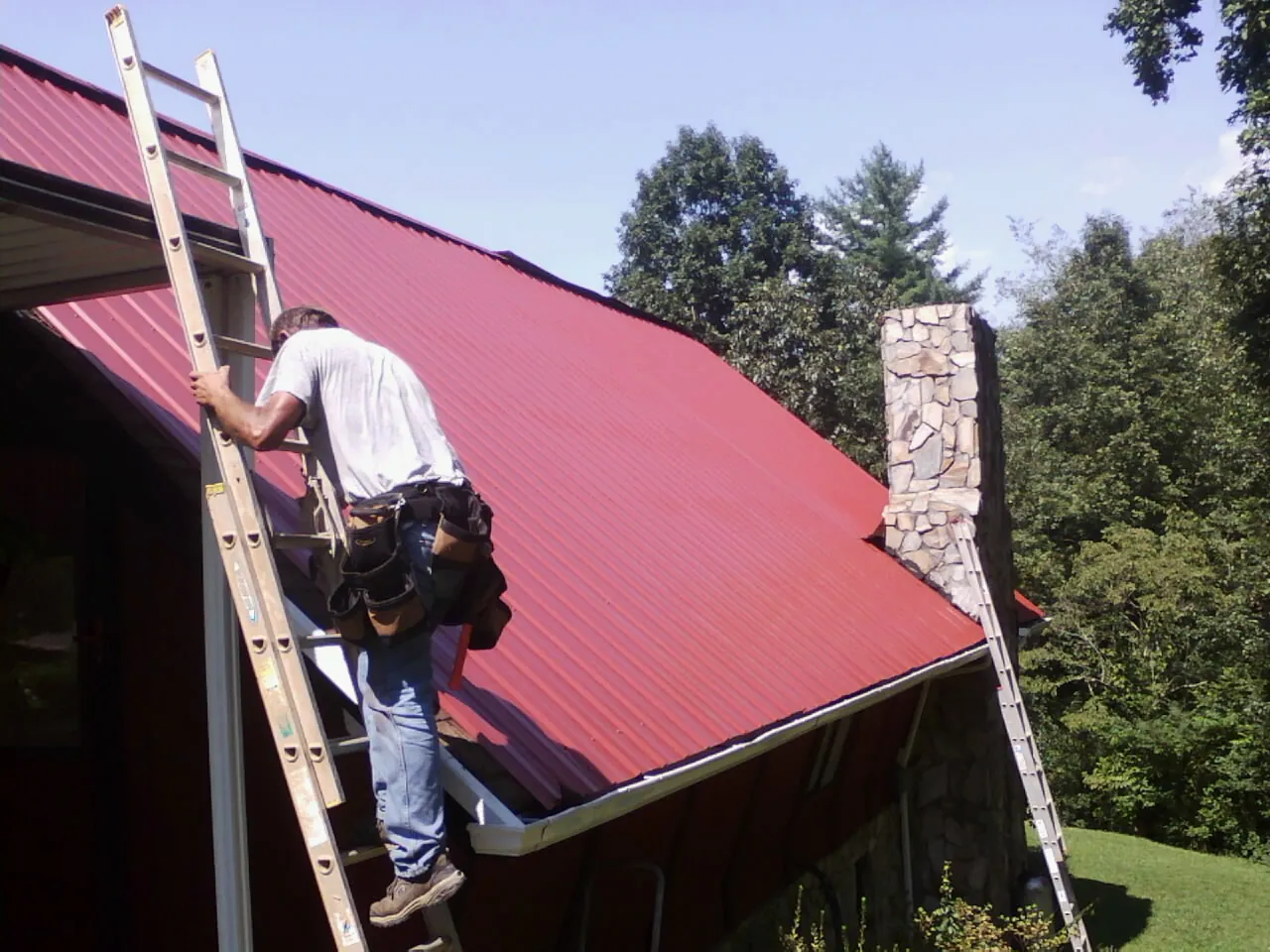 Roofer on a ladder beside a red metal roof and stone chimney — Soffit & Fascia Repair by Roofing Brothers in Enfield